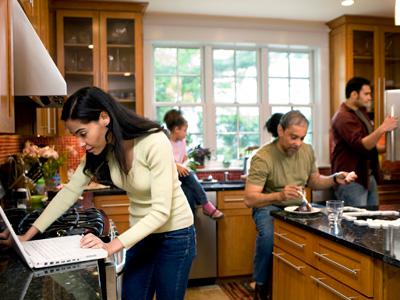 Busy family in a kitchen. Mom on laptop, dad eating, kids in background.